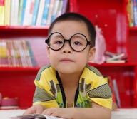 Young child with glasses in front of red painted shelves, leaning on table with open book in hand.